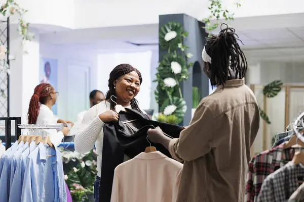 Smiling couple checking jacket fabric quality while shopping in fashion boutique. African american woman showing outfit on hangers to man and asking for advice before buying