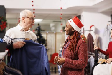 Alışveriş merkezi çalışanı müşteriye giysilerini gösteriyor, Noel yemeği için mükemmel ceketi seçmesine yardım ediyor. Afrikalı Amerikalı kadın çalışan kıdemli müşteriler için kıyafet öneriyor.