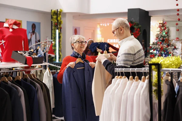 Joyful old people comparing items in store, showing each other trendy jackets to buy for festive christmas dinner. Senior clients smiling while shopping for clothes on discount, xmas decor.