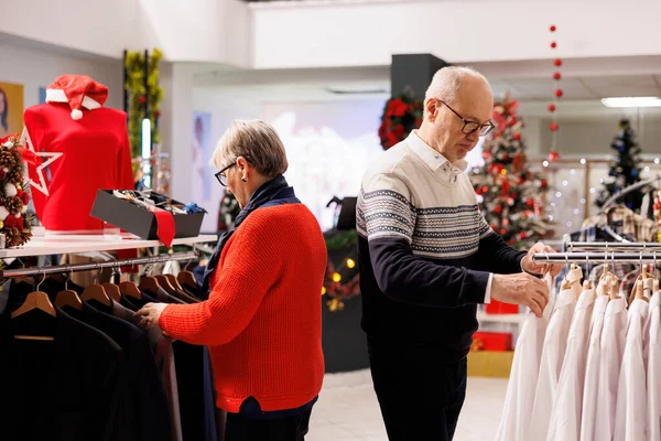 Elderly people searching for clothes in shopping center, looking at fashion items on hangers during winter season. Couple checking new merchandise in retail store, buying presents for family.