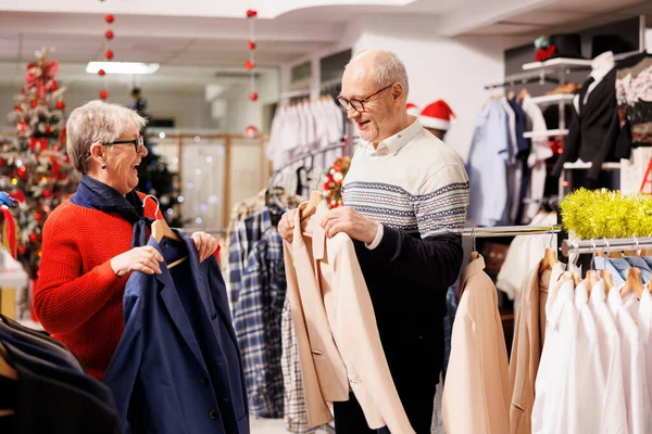 Senior people searching for jackets in retail store with festive ornaments, buying clothes as presents for family on christmas eve. Cheerful couple browsing through merchandise on sale.