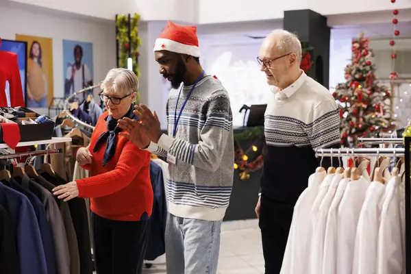 Employee showing items to senior couple shopping for family gifts, looking for presents in festive clothing store. African american retail assistant helping elderly people to choose perfect clothing.