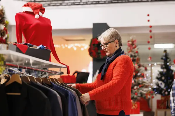 Senior client searching for clothes in fashion store during seasonal promotions, looking for Christmas dinner attire in boutique with festive decor. Woman customer at shopping center buys gifts.