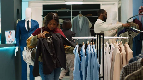 アフリカ系アメリカ人女性はハンガーから正装して衣料品店でワードローブを増やす 若いクライアントは ブティックで生地を分析し 新しい服コレクションを購入しようとしています 手持ち撮影 — ストック写真