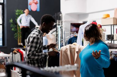 Moda koleksiyonu için şık bir müşteri alışverişi, modern butikte gömlek kumaşına bakmak. Afrikalı Amerikalı çift şık kıyafetler alıyor, elbise mağazasında resmi giysileri olan askıları kontrol ediyor.
