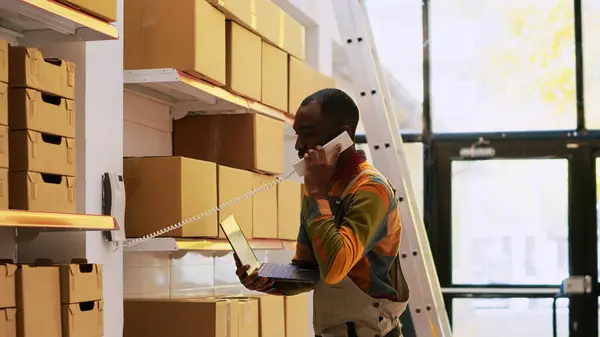 African american man inspecting products and talking about distribution with headquarters, cargo stock. Male supervisor using landline phone in storage room, quality control verification.