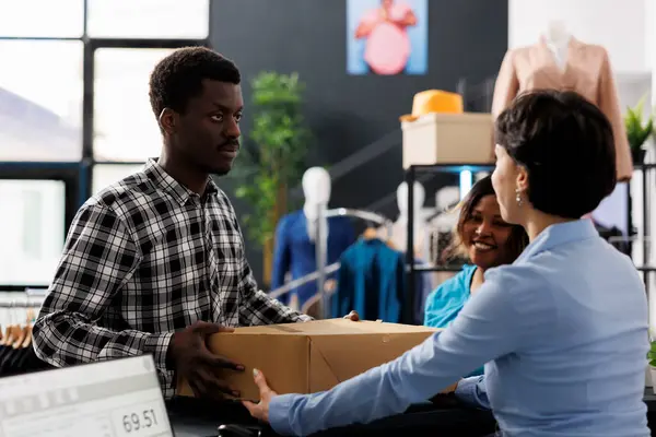 Clothing store employee giving online order to african american customer, standing at counter desk in modern boutique. Shopaholic man buying fashionable merchandise in shopping mall