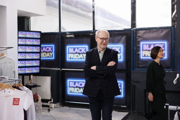 Friendly senior retail business owner wearing formal suit standing in clothing store against Black Friday neon banners, smiling man greeting and inviting customers to buy clothes during seasonal sales