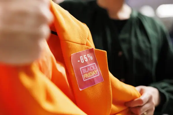 Black Friday fashion deals. Close up of red discount sale tag on clothing item. People buying clothes at discounted prices in shopping mall during seasonal sales, shoppers fighting for orange blazer