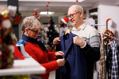 Perakende mağazasında spor ceketlere bakan yaşlı bir çift Noel yemeği kutlamalarında giymek için resmi kıyafetler arıyor. Müşteriler şık kıyafetler, şenlikli dekorlar için takım elbise kumaşlarını kontrol ediyor..