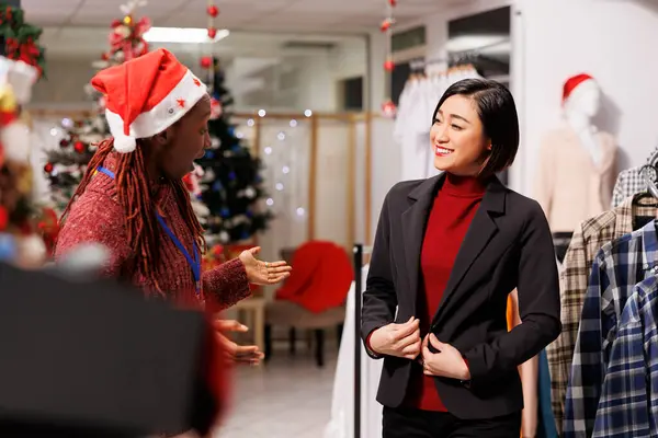 亚洲客户在购买节日服装前试穿外套 员工们帮助女人选择服装与家人共进圣诞大餐 向客户推荐西服的非裔美国人助理 — 图库照片