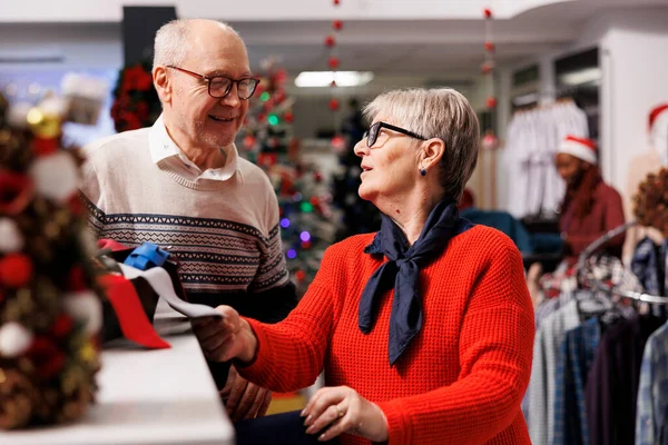 Cheerful elderly couple shopping at mall, looking to buy formal attire for christmas eve dinner outfit. Senior people checking ties from accessories box at clothing store, xmas decorations.