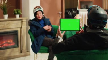 Genç adam, elinde boş krom anahtar modelleme şablonu olan dijital bir cihaz tutarak kayak tesisi alanındaki yeşil ekran ekranlı tablet kullanıyor. Çiftler kış sporları hakkında konuşuyor..