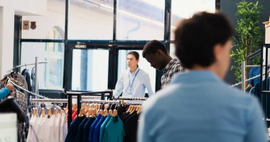 Afro-amerikan müşteriler modaya uygun gömlekli askılara bakıyorlar, giysileri almadan önce analiz ediyorlar. Alışveriş manyağı adam modern butikte şık ürünler ve aksesuarlar alıyor.