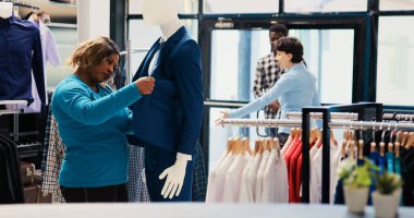 Afro-amerikan müşteri şık bir takım elbiseye bakıyor, modern butikte kumaş ürünlerini analiz ediyor. Alışverişkolik kadın şık kıyafetler alıyor, giyim mağazasından moda eşyalar alıyor.