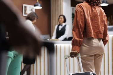 Woman waiting to do check in at hotel, carrying luggage at reception area with front desk. Young female guest preparing to register for room accommodation at luxury resort, lobby space. clipart