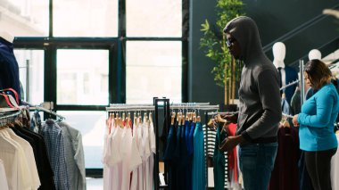 Afrikalı Amerikalı hırsız şık gömlekler çalıyor, modern butikten kaçmaya çalışıyor. Asyalı koruma mağaza kapısında soyguncuyu yakalayıp moda kıyafetleri iade etmesini yoksa polisi arayacağını söyledi.