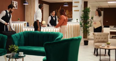 Dostça gülümseyen kapıcı personel otel lobisinde misafirleri karşılıyor. Profesyonel komi, turistlerin bagajlarını taşımalarına yardım ederken neşeli resepsiyonist de yardım çeki veriyor.