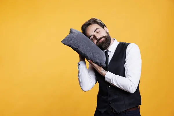 Sleepy butler sleeping on pillow, taking a nap while he in standing in studio. Young exhausted overworked person falling asleep against yellow background, working in restaurant industry.