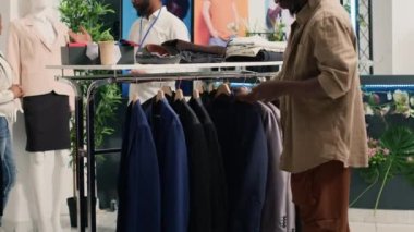 Giyim mağazasında şık ceketlere bakan bir adam malzemelerin nitelikli olup olmadığını belirlemeye çalışıyor. Moda mağazalarında giysi analizi yapan müşteriler, en iyi kaliteyi bulduktan sonra almaya karar verirler.