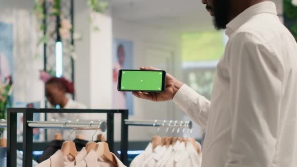 エレガントなフォーマルウェアシャツで高級衣料品店で緑色のスクリーン携帯電話を持っているアフリカ系アメリカ人男性 ファッションブティックでクロマキー携帯電話を使用したBipoc顧客 — ストック動画