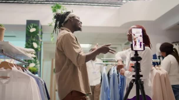 互联网明星旁边的商店员工使用智能手机拍摄服装商店与优雅的服装组合 有影响力的人花钱向追随者展示新的服装系列 — 图库视频影像