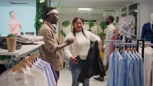 アフリカ系アメリカ人男性と彼のガールフレンドは 醜いブレザーに不満を持って第二の手の衣料品店で安い服を探しています 彼女のボーイフレンドとディスカウントストアで写真を撮ろうとしている女性 — ストック動画