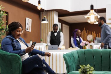 Mutlu, rahat, Afro-Amerikan kadın otel lobisinde akıllı telefon kullanarak internet ya da internet izleyerek rahatlıyor. Lüks tatil beldesindeki dinlenme yerinde telefonla dinlenen kadın turist.