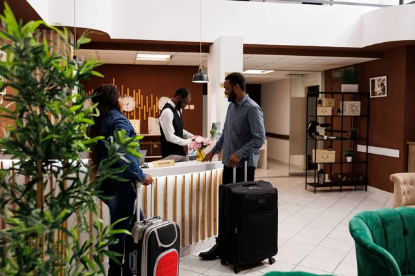Happy black couple standing at hotel front desk with luggage, checking in at honeymoon resort, people waiting at reception for check-in. Receptionist helping guests with check out