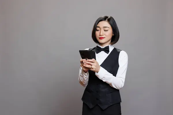 Smiling asian restaurant waitress in black and white uniform typing message on smartphone. Catering service attractive woman employee using mobile phone while posing in studio