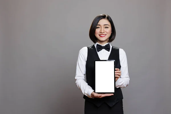 Restaurant asian woman receptionist holding digital tablet with white empty screen for catering service app. Smiling cheerful waitress in unform showing blank touchscreen for promotion