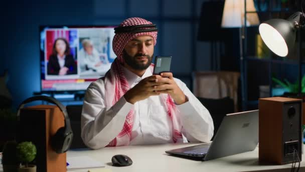 スマートフォンでメッセージを入力するリビングルームのアラビア人男性 携帯電話を持っているイスラム教徒は オンラインメッセージングアプリで友達と話すリラックスしたレジャータイムを楽しんでいます — ストック動画