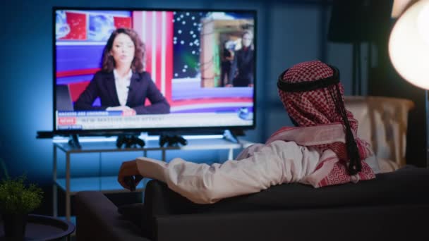 アラブの男は居心地の良いソファーにぶら下がり 仕事で忙しい一日の後に自宅でリラックスしながらニュース放送を見ました イスラム教徒はソファーで冷やし 現在のイベントを示すテレビチャンネルを見て — ストック動画
