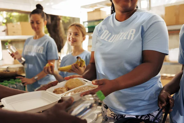 At non-profit food bank, african american woman serves baked beans to the less fortunate individual. Close-up of humanitarian aid female team distributing free warm meals and nourishments to the needy
