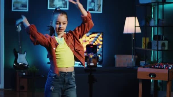 素敵なダンスの動きをしている小さな女の子は スマートフォンを使ってウイルス上の挑戦を撮影します 薄暗く照らされた家のスタジオのオンライン ファンのための携帯電話が付いている楽しい録画ビデオを持っている幸せな子供 — ストック動画