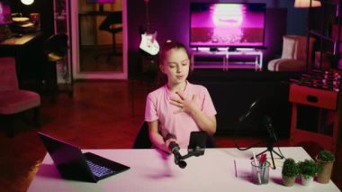 Oturma odasında sosyal medya platformu için video çeken mutlu bir kızın gününden bahsettiği bir kare. Neşeli çocuk hayranlarını eğlendiriyor, selfie çubuğunda telefon kullanarak içerik yaratıyor.