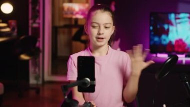 Profesyonel stüdyo olarak kullanılan pembe neon ışıklı oturma odasında selfie çubuğuna bağlı akıllı telefonlu çocuk filmleri. Genç medya yıldızı cep telefonu kamerasıyla okulda eğlenceli bir gün hakkında konuşurken görüntüler yakalıyor.