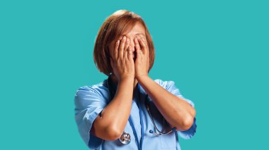 Bitkin düşmüş BIPOC maharetli hemşire avuçlarıyla yüzünü kapatıyor, uzun iş vardiyasından sonra uykulu. Asyalı sağlık çalışanı işinden ayrılmaya ihtiyacı var. Stüdyonun arka planında izole edilmiş, kapatılmış.