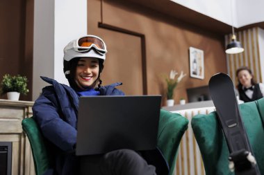 Kışlık kıyafetler giyen Asyalı kadın turist otel lobisindeki dijital cihazla kanepenin keyfini çıkarıyor. Kayak ekipmanlarıyla heyecanlı bir kadın laptopta kayak merkezinde yamaç arıyor..