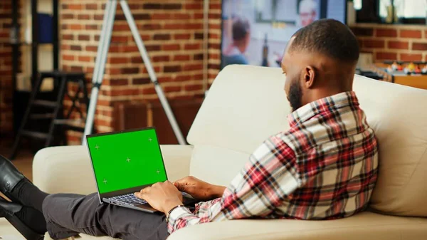 Focused remote freelancer working from home, typing data on chroma key mock up laptop green screen. African american teleworker solving company tasks in cozy apartment living room