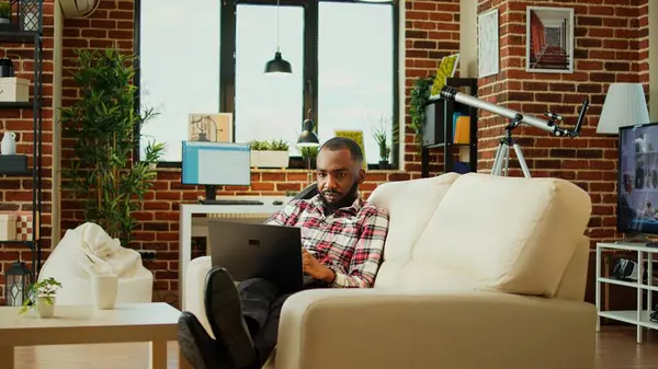 Relaxed freelancer inputing data on laptop while working from home. African american teleworker lying on couch, remotely performing tasks in nice stylish warm apartment living room