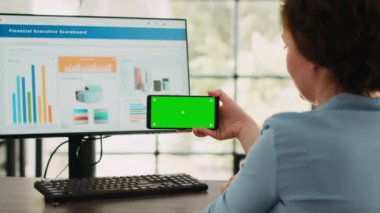 İşletme uzmanı akıllı telefon yazılım ekranında yeşil ekran gösteriyor, izole edilmiş kromakey şablonuna bakıyor ve masadaki bilgisayarda çalışıyor. Boş maketler kullanarak ve teşkilat operasyonlarını tamamlayarak..