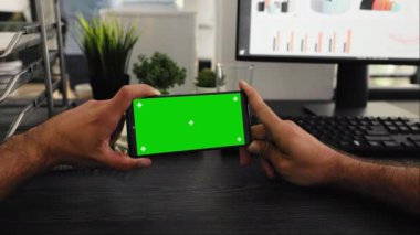 Çalışma masasında yeşil ekran görüntülü POV, çalışma istasyonunda izole modelleme şablonu ile çalışıyor. Genç çalışan, krom anahtarlı cep telefonu ekranına bakıyor.