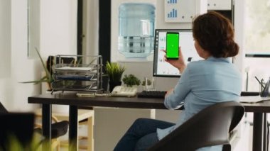 Çalışan akıllı telefonu elinde tutuyor ve küçük işletme ofisinde yeşil ekran ekranı gösteriyor. Ekran düzeninde izole edilmiş kopyalama alanı olan mobil aygıta bakan genç bir kadın.