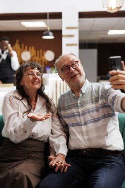 Lüks otel lobisinde, emekli üst düzey müşterilerin dinlenme odasında online video görüşmeleri yaparken detaylı görüntüsü. Balayındaki mutlu yaşlı çift, cep telefonuyla aileleri ve arkadaşlarıyla iletişim kuruyor..