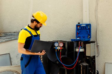 Açık hava klima iç bileşenlerini tamir etmekle görevli eğitimli bir mühendis, freon seviyesini kontrol ediyor ve termostatları ayarlıyor. Dizüstü bilgisayarda bakım planına bakan yetenekli bir teknisyen.