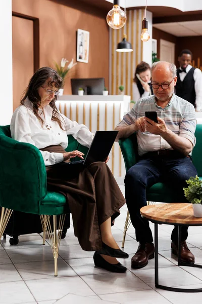Caucasian old man using mobile phone while elderly female tourist browsing the internet on her personal computer. Retired senior couple sitting in lounge area surfing the net on their digital devices.