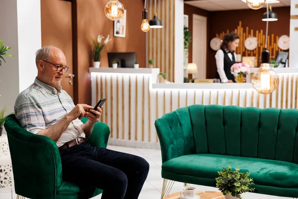 stock image Elderly male customer seated on sofa using mobile device while waiting for check-in in hotel lobby. Retired senior man surfing the net for holiday activities on his smartphone in lounge area.