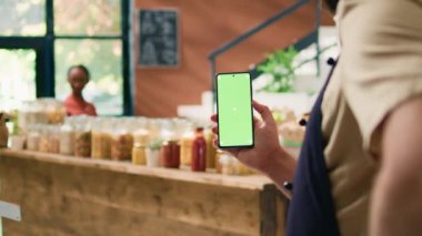 Yerel satıcı akıllı telefon uygulamasını yeşil ekran ekranlı tutuyor, mobil cihazda telif alanı teknolojisi sunuyor. Genç adam manavdaki kasada izole edilmiş bir model ekranı gösteriyor..