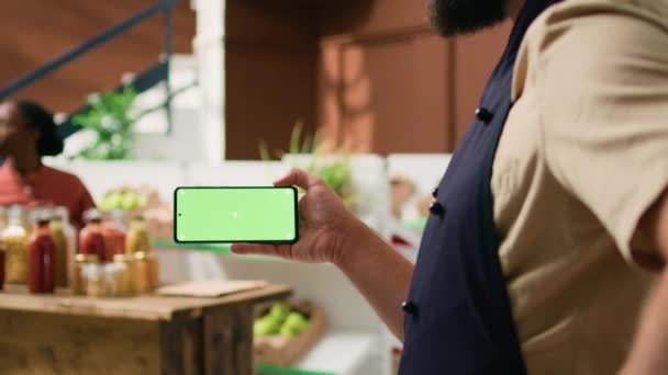 地元のベンダーは 単離ディスプレイ付きのグリーンスクリーン携帯電話を保持し 携帯電話でコピースペース技術を提示しています 食料品店のレジで空白クロメイクスクリーンを見ている人 — ストック動画